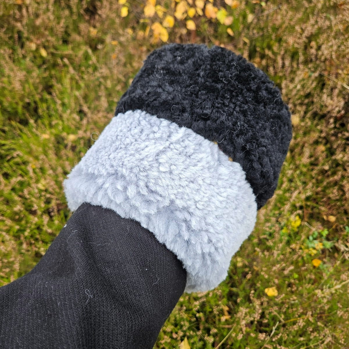 Hand som håller ett svart och grått fårskinnspannband utomhus i naturmiljö med höstfärger i bakgrunden.