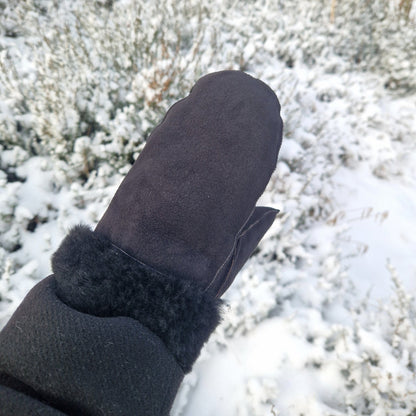 Hand som bär svart fårskinnsvante med svart ullfoder, fotograferad utomhus i vintrigt snölandskap – varma och mjuka handgjorda vintervantar i äkta fårskinn.